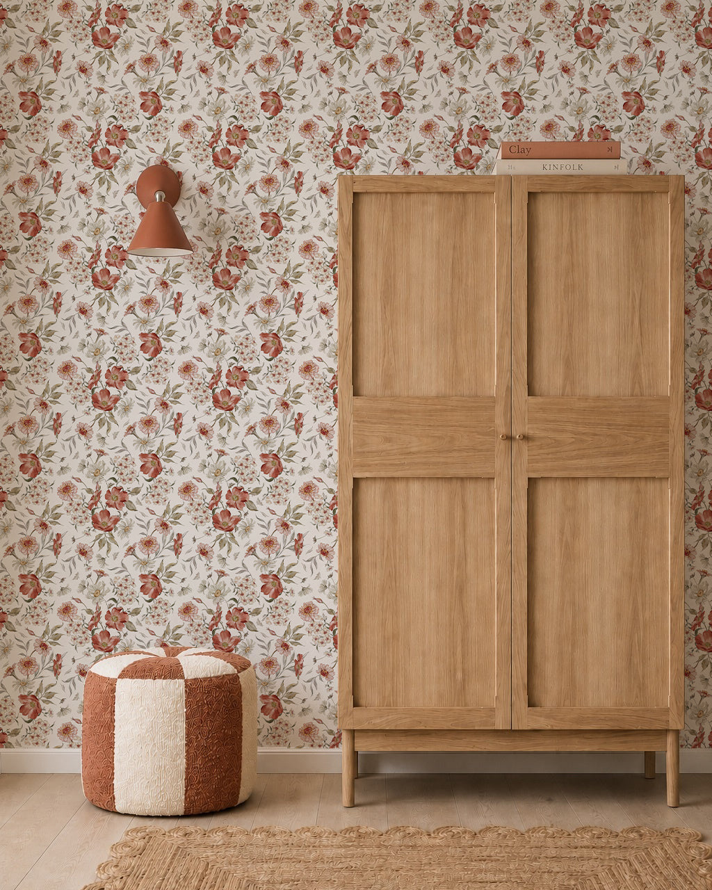A wooden wardrobe stands next to a red and cream pouf on a tan woven rug, with Wild Rose Dreams Wallpaper, a wall lamp, and books stacked on top of the wardrobe.