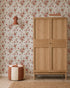 A wooden wardrobe stands next to a red and cream pouf on a tan woven rug, with Wild Rose Dreams Wallpaper, a wall lamp, and books stacked on top of the wardrobe.
