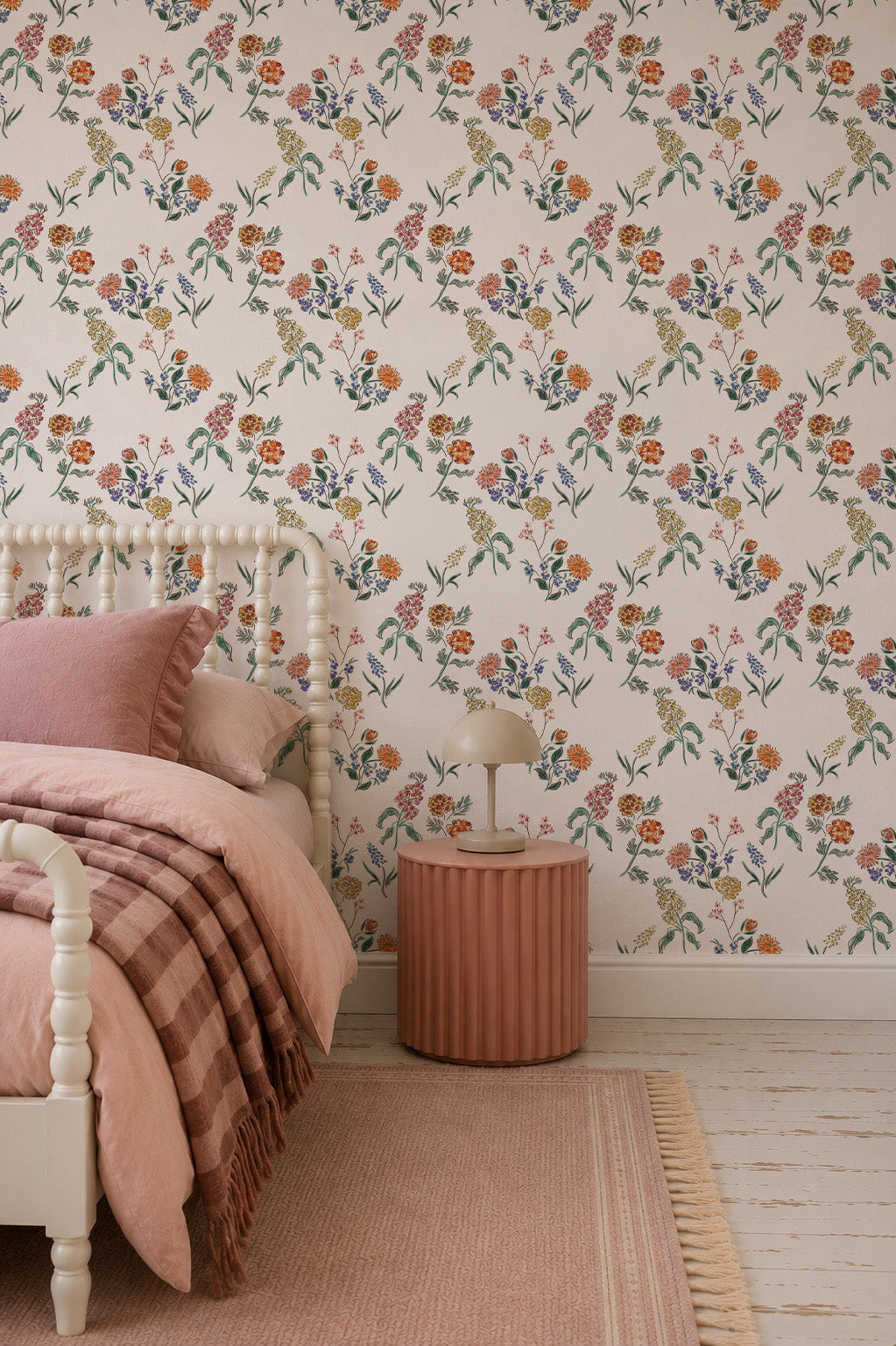 A cozy bedroom with Wildflower Posy Wallpaper featuring delicate floral patterns, pale pink bedding, a round pink nightstand with a small lamp, and a matching rug on a light wooden floor.