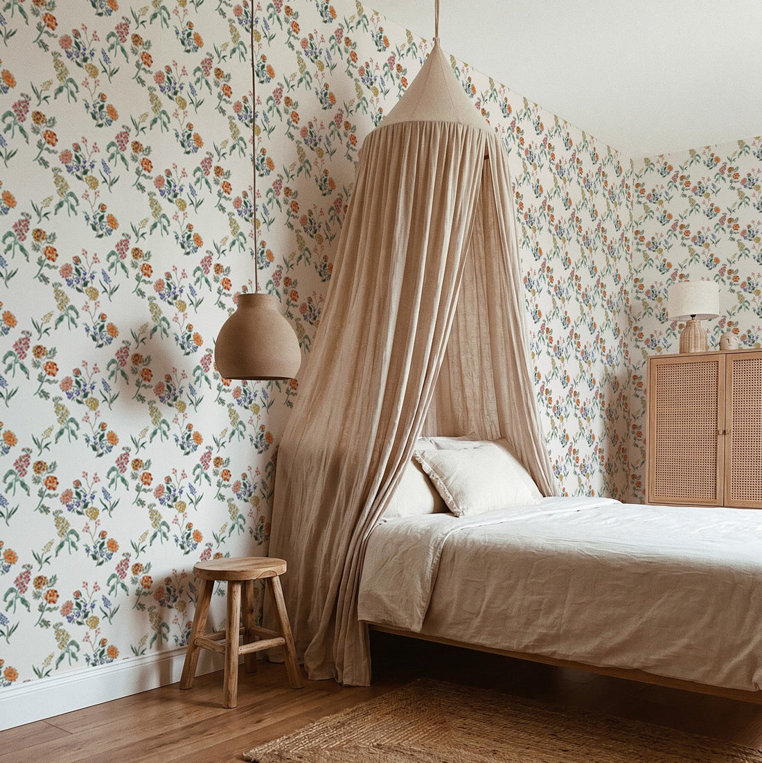 A cozy bedroom featuring Wildflower Posy Wallpaper, a canopy bed with beige bedding, a small wooden stool, and a rattan cabinet with a lamp. Warm, neutral tones create a calm and inviting atmosphere.