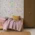 A cozy bedroom with a wood bed, purple checkered bedding, yellow pillow, and the Wildflower Waltz Wallpaper that brings a touch of nature decor. The room features a brown wardrobe and a woven rug on white wooden floors.