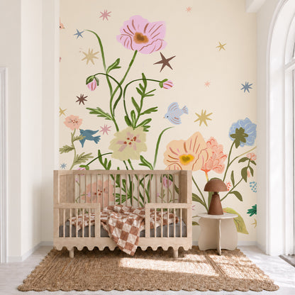 A wooden crib, The Wishing Garden, Cream, sits in front of a wall featuring a whimsical pink floral wall mural decorated with stars and small birds.