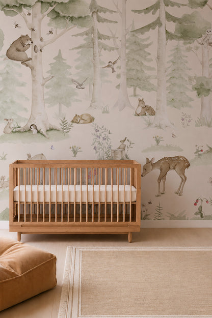 A wooden crib sits in a nursery adorned with the Woodland Friends Mural, which showcases deer, bears, and birds. A tan ottoman and beige rug rest on the light wood floor, creating a calm, nature-inspired atmosphere.