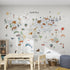 Children’s playroom with a cozy setup featuring the World Map Mural, Grey—an educational wallpaper illustrated with animals and landmarks. A small table, rug, toys, pouf, and shelves complete this inviting kids’ room.