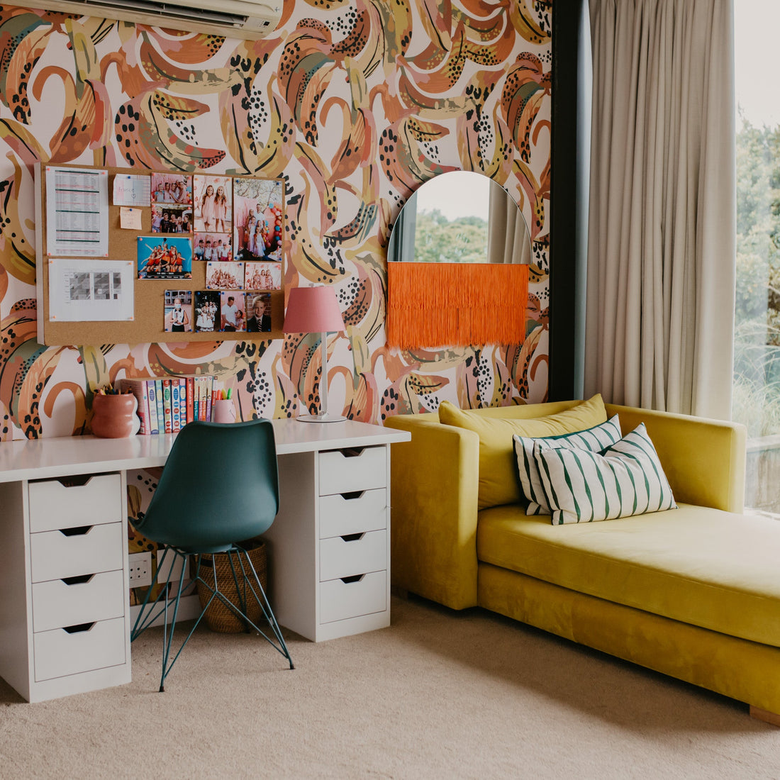 A colorful room features Banana Party Wallpaper, two green chairs at a white desk, photos on the wall, and a vibrant yellow chaise lounge by large windows overlooking green trees.